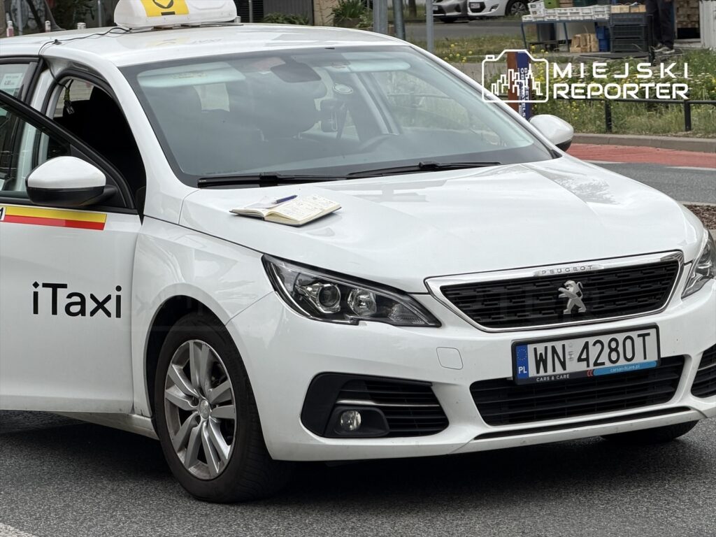 Biały taksówka marki Peugeot stoi na ulicy, z otwartymi drzwiami i notatnikiem na dachu.
