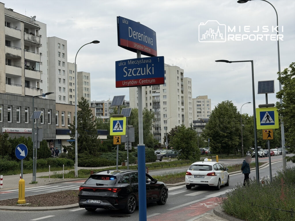 Ulica z nowoczesnymi budynkami mieszkalnymi, znakami drogowymi oraz taksówką zatrzymującą się przy przejściu dla pieszych.