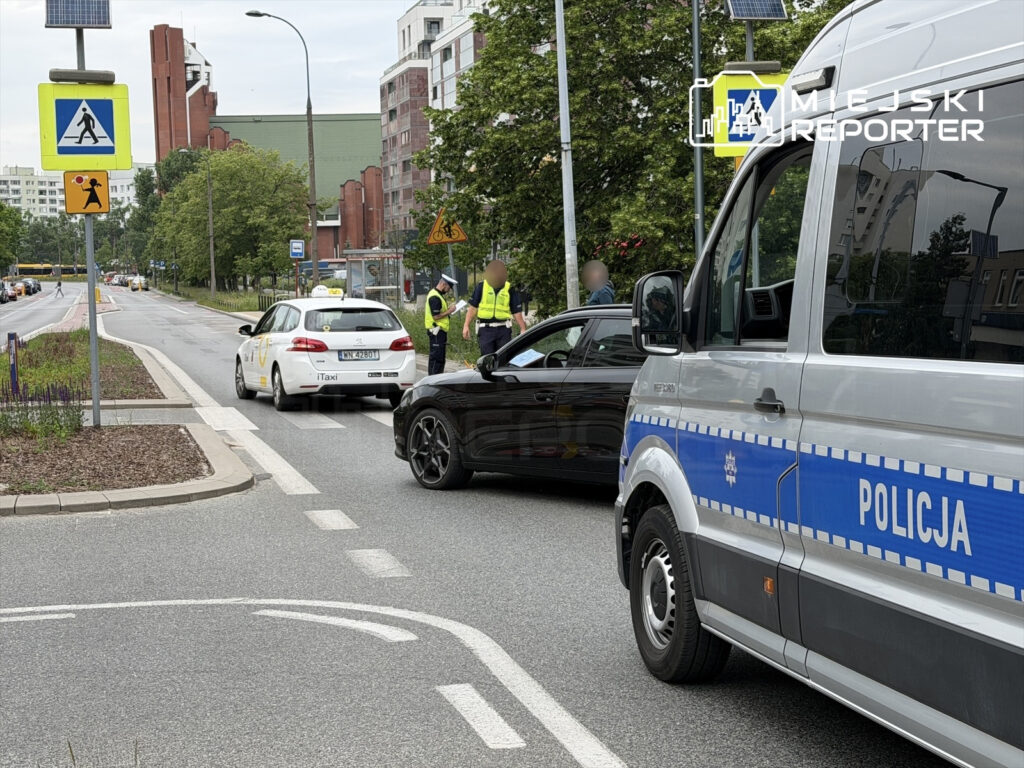 Policjanci kontrolują taksówkę na ulicy, obok zatrzymanego czarnego samochodu, w otoczeniu zieleni i budynków mieszkalnych.