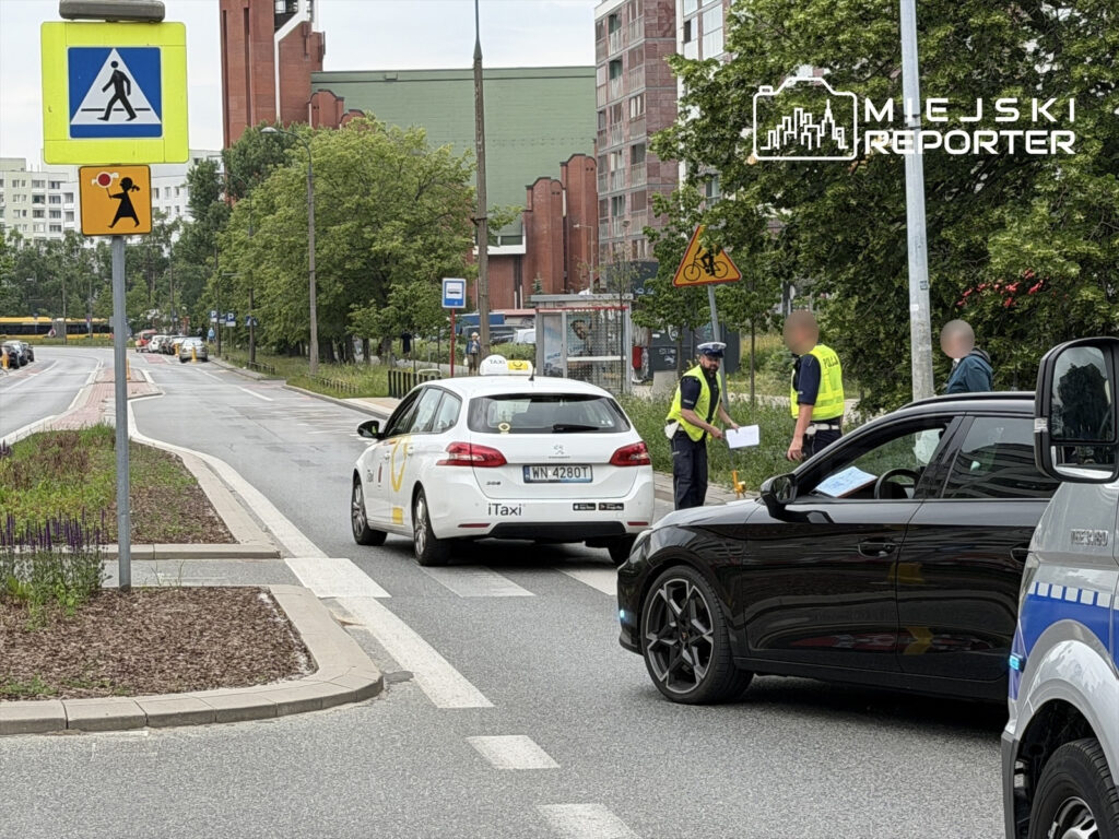 Policjanci kontrolujący kierowców na ulicy, obok taksówki i samochodu osobowego, w tle znaki drogowe.