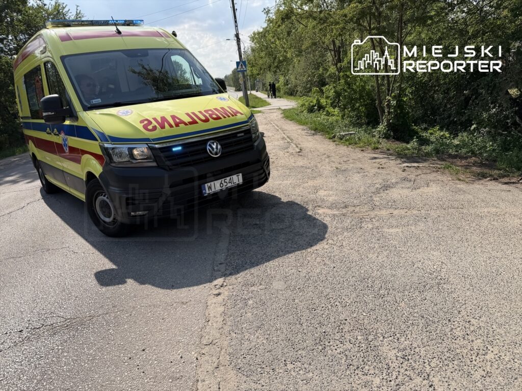 Ambulans w kolorach służb ratunkowych zatrzymany na poboczu drogi, w tle zieleń drzew i krzewów.