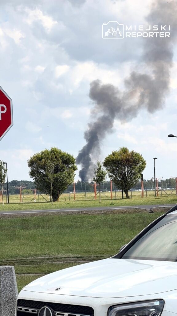 Czarny dym unoszący się z daleka, w tle zielona przestrzeń i znaki drogowe, w tym znak STOP.