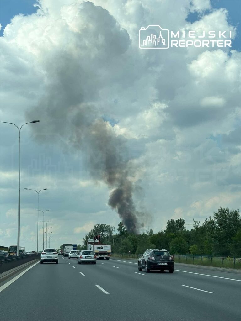 Na drodze widoczny jest dym unoszący się z ognia wśród drzew, obok poruszające się samochody.