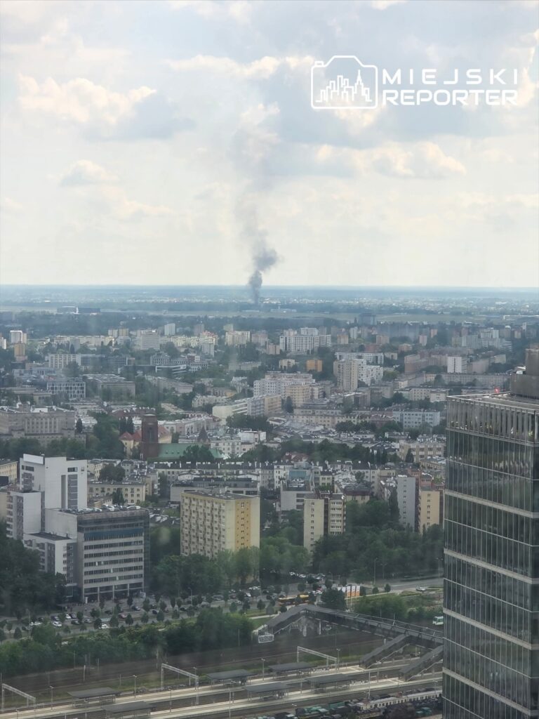 W tle widoczny dym unoszący się nad miastem, w otoczeniu nowoczesnych budynków i zieleni.
