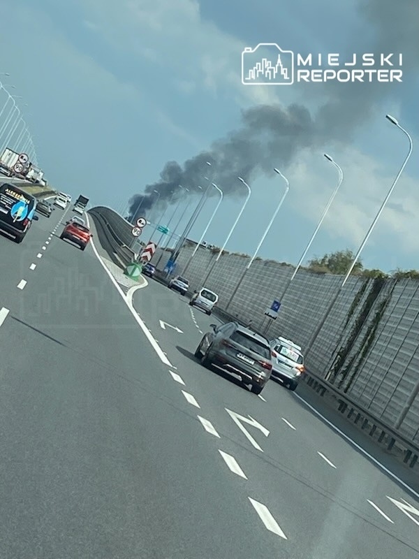 Na autostradzie widać dym unoszący się z daleka, a samochody poruszają się w różnych kierunkach.