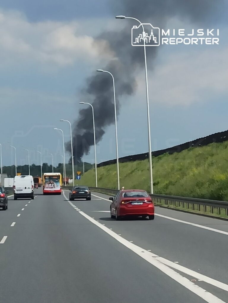 Czarny dym unoszący się z daleka nad autostradą, w otoczeniu samochodów osobowych i ciężarowych.