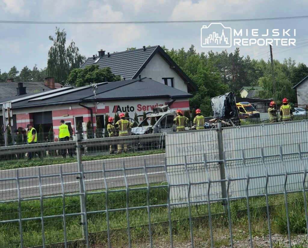 Grupa strażaków i policjantów pracuje przy uszkodzonym pojeździe obok warsztatu samochodowego.