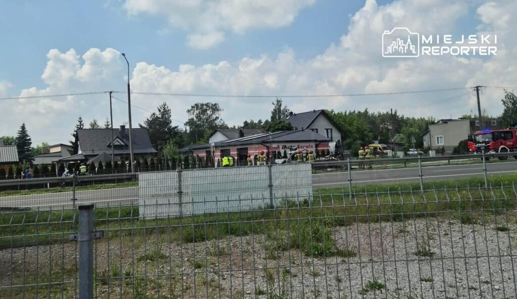 Grupa ratowników pracuje przy wypadku drogowym obok serwisu samochodowego, w tle widoczne domy i niebo z chmurami.