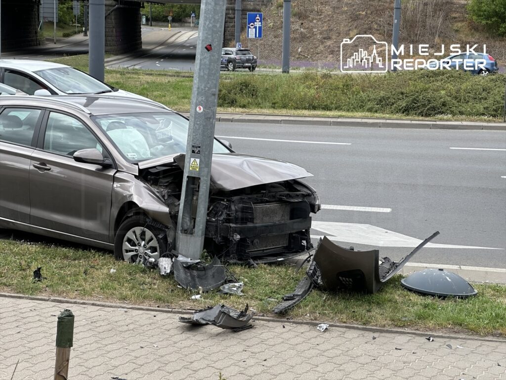 Uszkodzony samochód osobowy uderzył w słup oświetleniowy na poboczu drogi, obok leżące fragmenty karoserii.