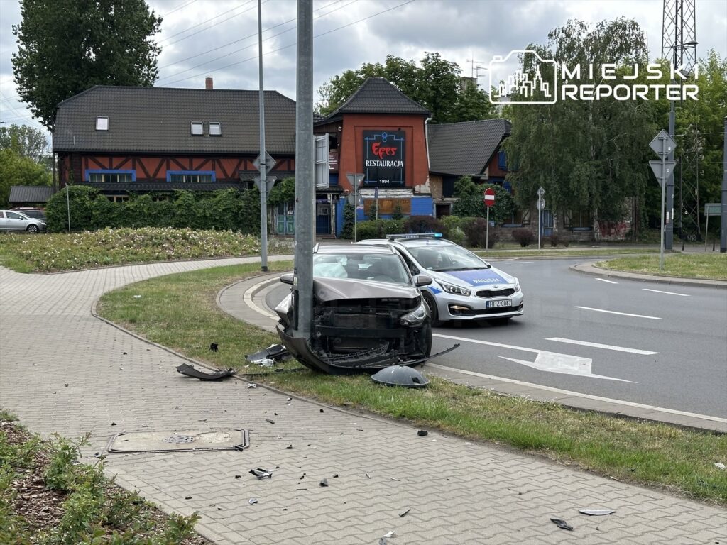 Zniszczony samochód osobowy stoi na poboczu przy rondzie, obok policyjny radiowóz. W tle widać budynek restauracji.