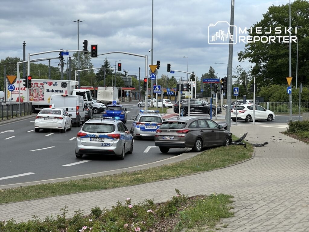 Na skrzyżowaniu ruchliwych dróg, kilka samochodów, w tym radiowozy, uczestniczy w akcji po kolizji, z jednym pojazdem uszkodzonym przy latarni.
