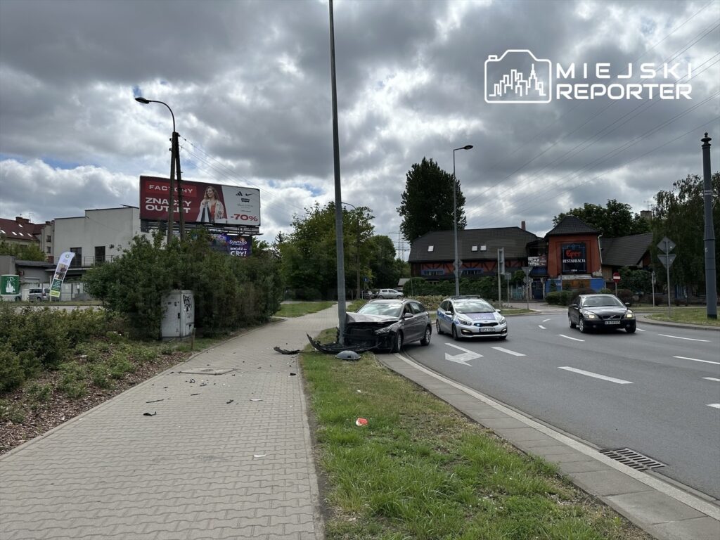 Samochód z uszkodzoną przednią częścią stoi na poboczu drogi, obok widać fragmenty wraku i billboard reklamowy.