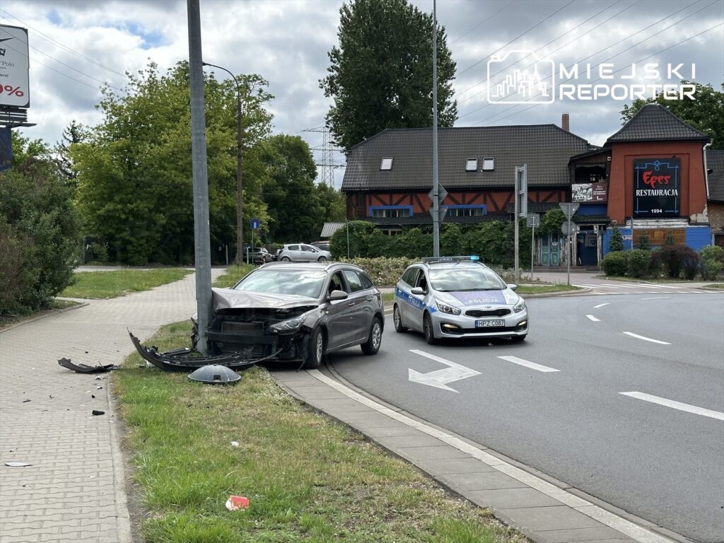 Uszkodzony samochód osobowy stoi przy latarni, obok policyjny radiowóz na skrzyżowaniu.