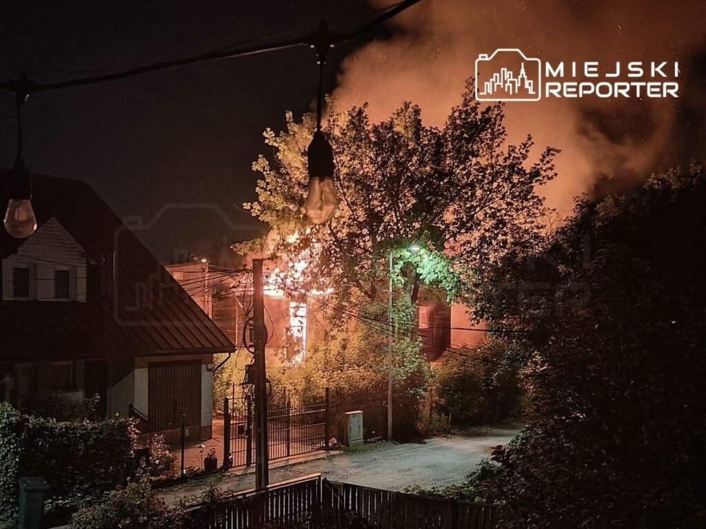 Ognisko pożaru w budynku mieszkalnym, otoczone drzewami i lampami ulicznymi, w ciemnej nocnej scenerii.