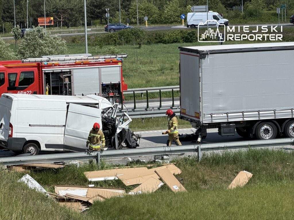Wielki pojazd dostawczy z uszkodzoną kabiną stoi przy drodze, a strażak w hełmie ocenia sytuację obok.