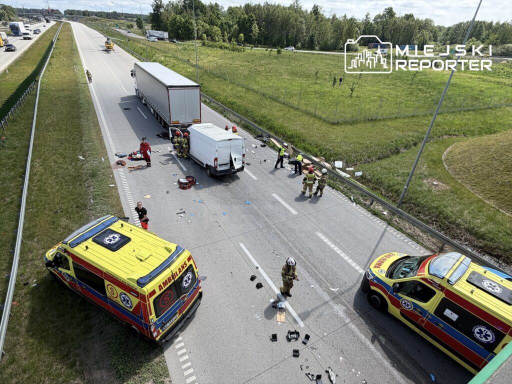 Na jezdni leżą resztki pojazdów po wypadku, a ratownicy medyczni i strażacy udzielają pomocy poszkodowanym.