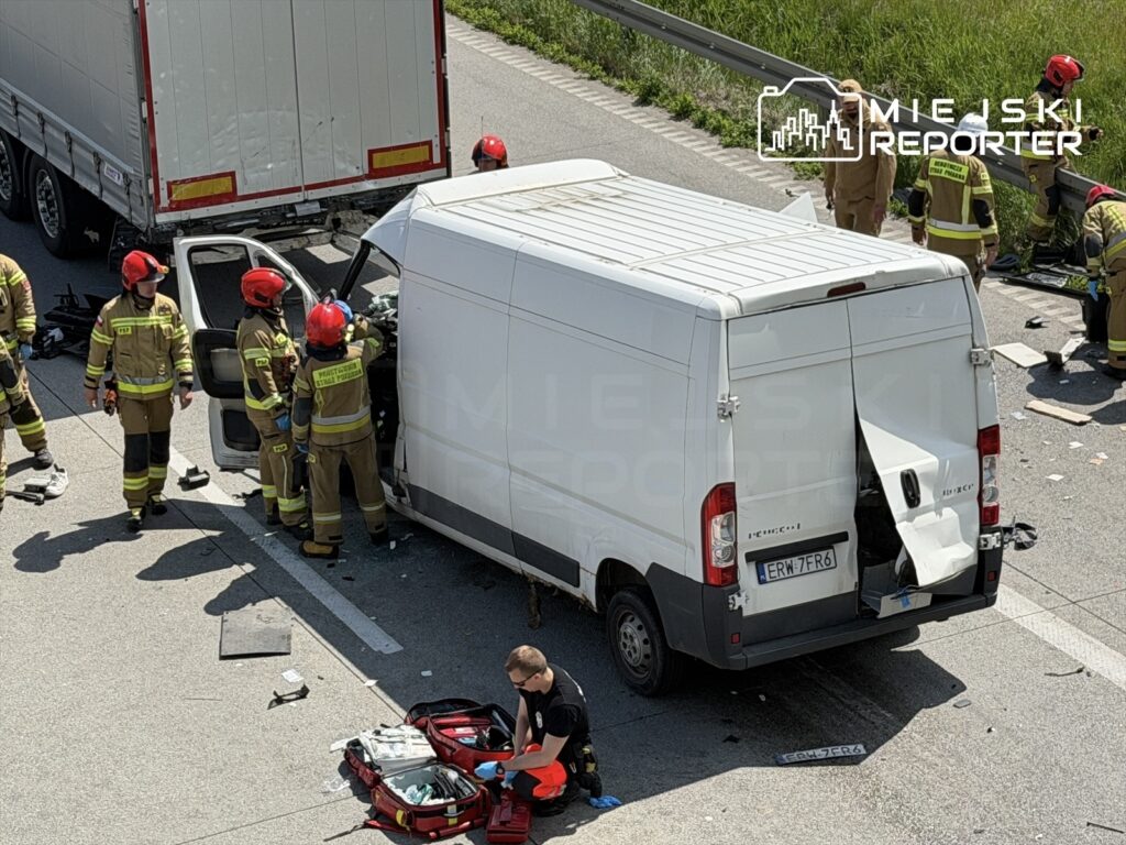 Grupa strażaków w hełmach bada uszkodzony dostawczy samochód na drodze, obok leżą sprzęt ratunkowy.