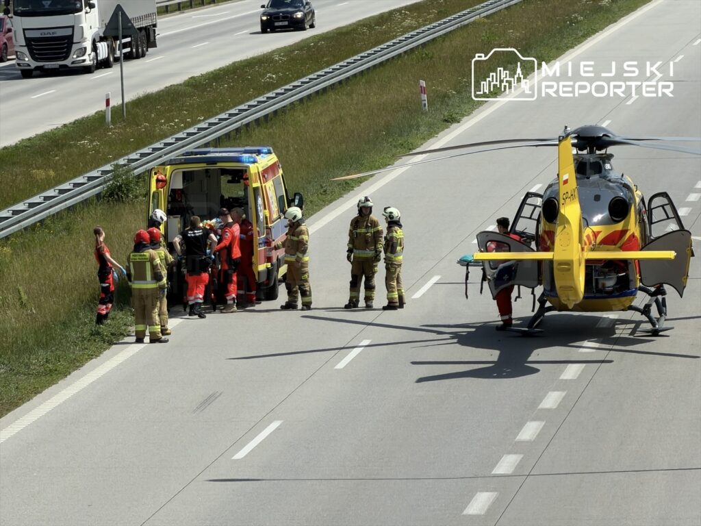 Grupa ratowników w czerwonych strojach udziela pomocy medycznej przy otwartej karetce na poboczu drogi, obok lądował śmigłowiec ratunkowy.