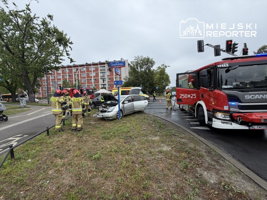 Grupa strażaków w mundurach interweniuje przy rozbitym samochodzie osobowym na ruchliwym skrzyżowaniu.