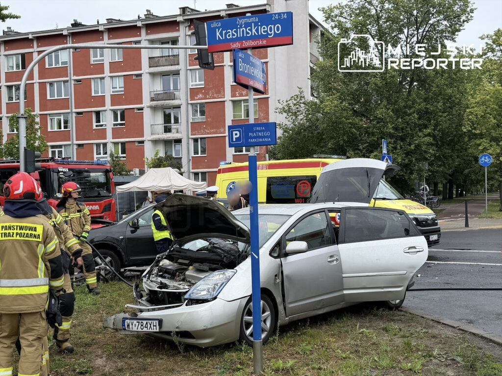 Grupa strażaków w mundurach pracuje przy uszkodzonym samochodzie osobowym w miejskim otoczeniu, obok widać wóz strażacki.