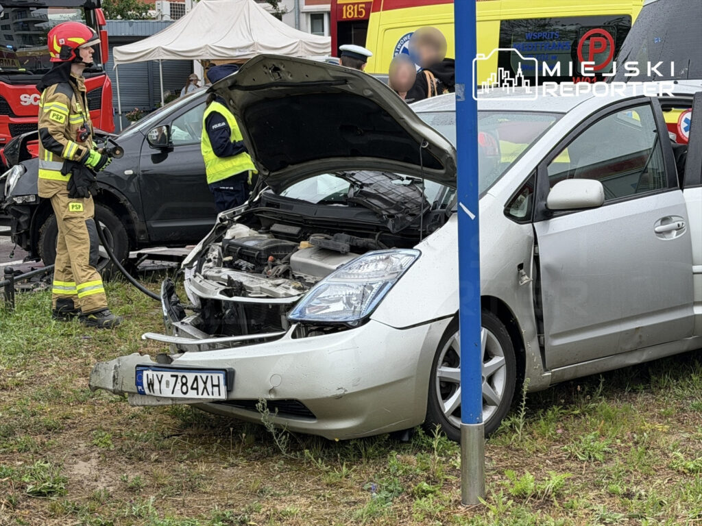 Strażak w mundurze trzymający sprzęt gaśniczy obok uszkodzonego samochodu osobowego na trawniku.