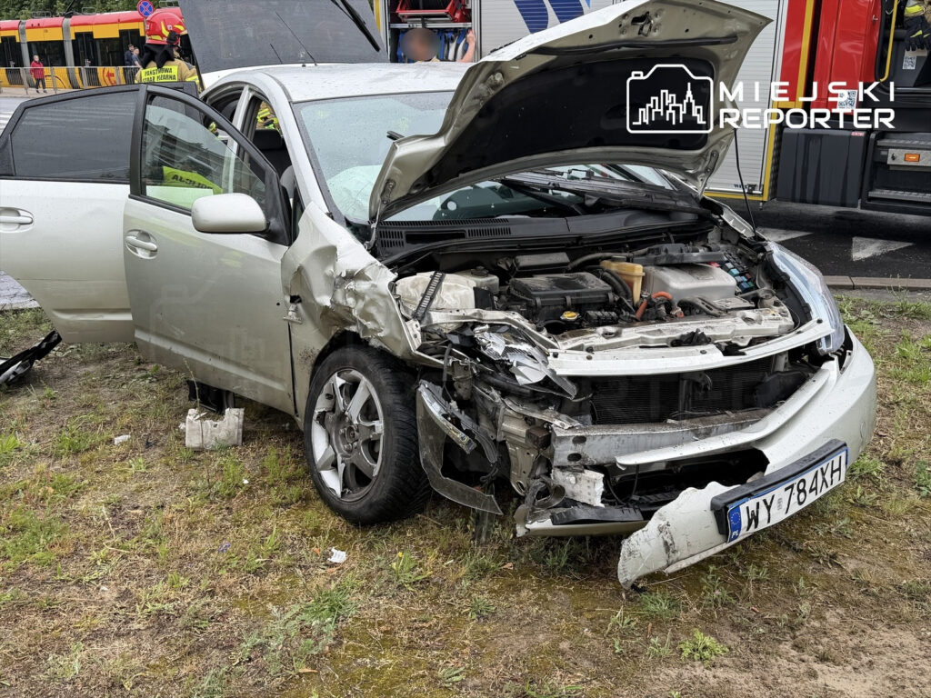 Uszkodzony samochód osobowy z wgniecioną przednią częścią stoi na trawie obok drogi, otwarta jest jedna z drzwi.