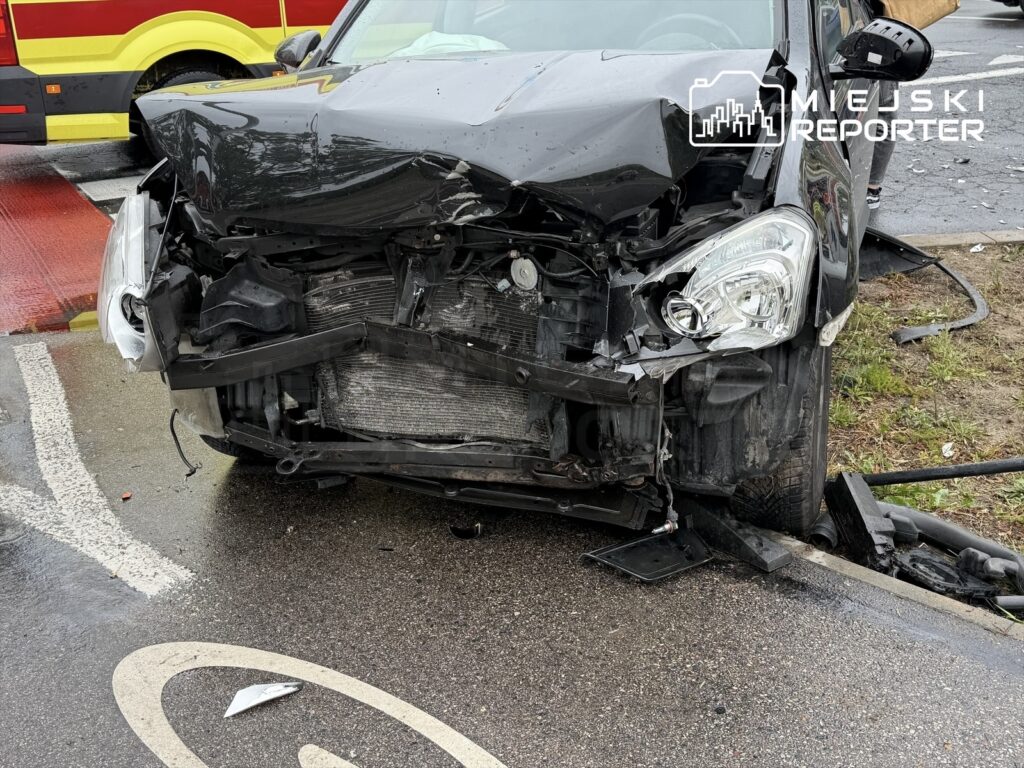 Zniszczony czarny samochód stojący na poboczu drogi, z uszkodzoną przednią częścią i widocznymi elementami silnika.