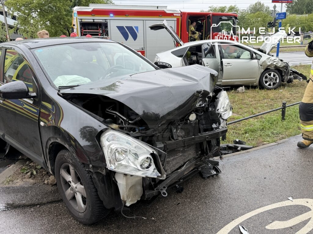 Czarny samochód osobowy z uszkodzoną przednią częścią stoi na poboczu drogi, obok widać uszkodzony srebrny pojazd oraz strażaków.