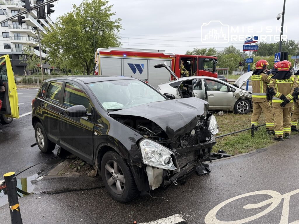 Czarny samochód osobowy z uszkodzoną przednią częścią stoi na poboczu, obok interweniujący strażacy w mundurach.