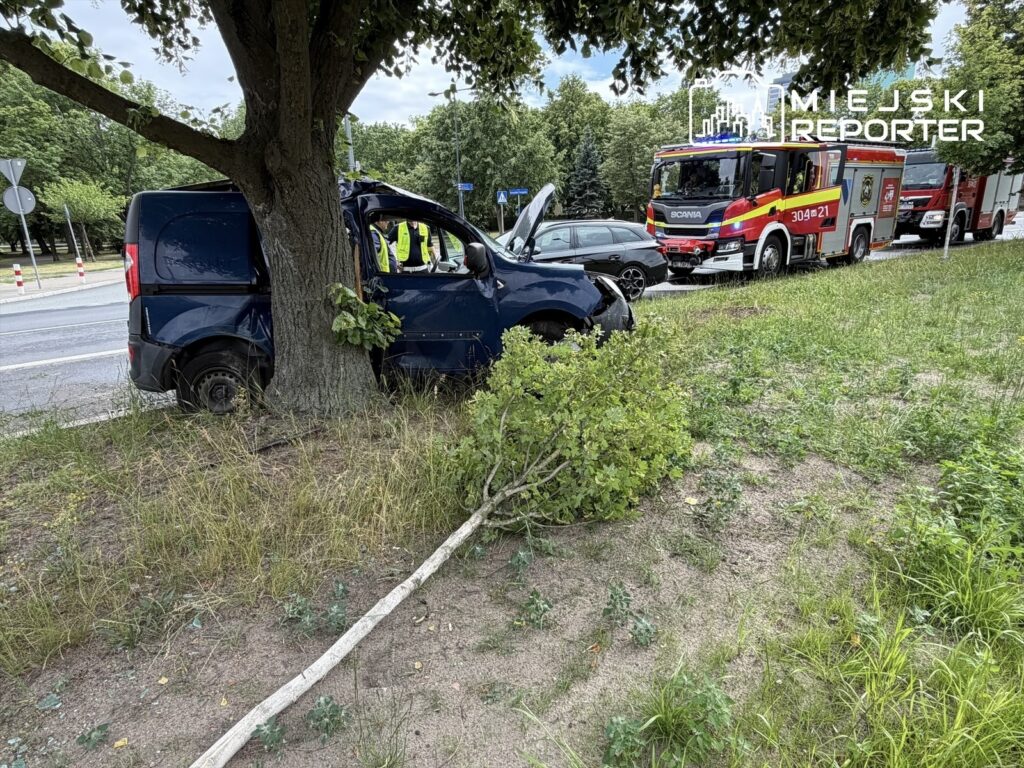 Niebieski samochód dostawczy uderzył w drzewo przy ruchliwej drodze, obok stoją wozu strażackiego.