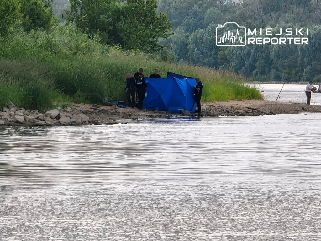 Grupa ratowników w strojach nurkowych ustawia niebieski namiot na brzegu rzeki, w tle widać wędkarza.