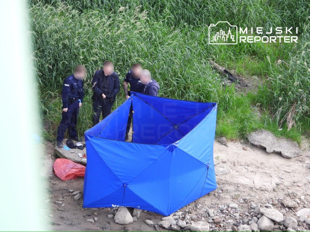 Grupa policjantów w mundurach stoi przy niebieskim namiocie na brzegu rzeki, otoczona zielonymi krzakami.