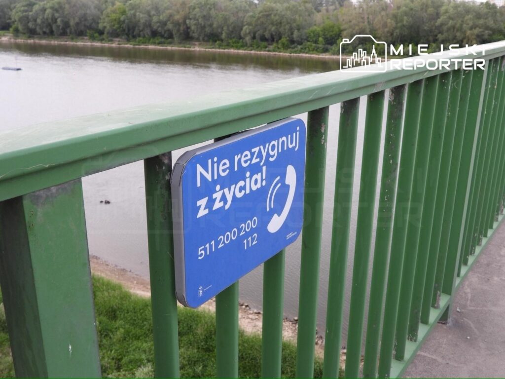 Zielona balustrada mostu z niebieskim znakiem informacyjnym "Nie rezygnuj z życia!" oraz numerami telefonów.