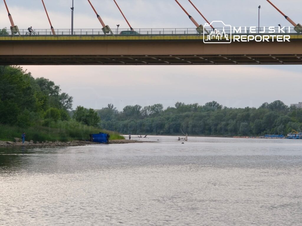 Rowerzysta przejeżdżający po moście nad rzeką, w tle zieleń i łodzie na wodzie.