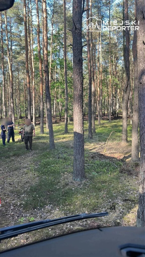 Grupa osób w mundurach rozmawia w lesie, otoczona wysokimi drzewami, na tle zielonej trawy i krzewów.