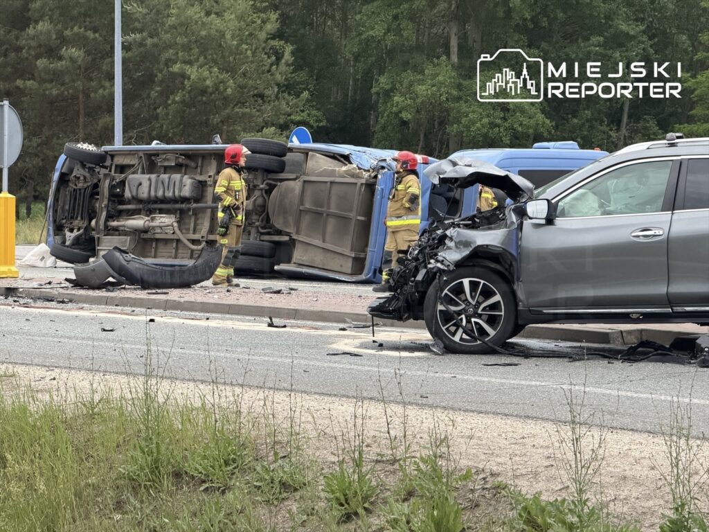 Strażacy pracują przy przewróconym samochodzie na drodze obok uszkodzonego pojazdu osobowego.