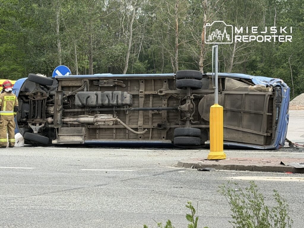 Nieoznakowany niebieski samochód leży na dachu na drodze, obok stojący strażak w mundurze i kasku.