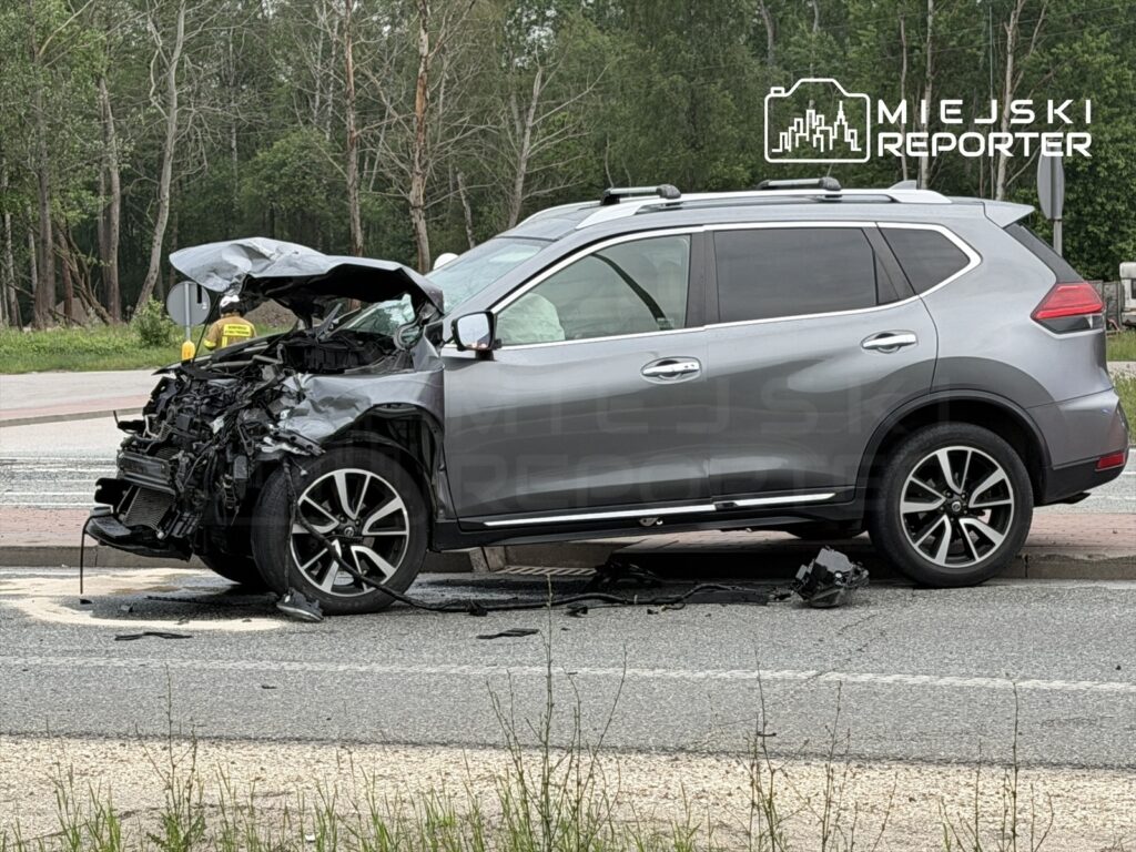 Rozbity samochód osobowy z uszkodzoną przednią częścią stoi na poboczu drogi w otoczeniu zieleni.