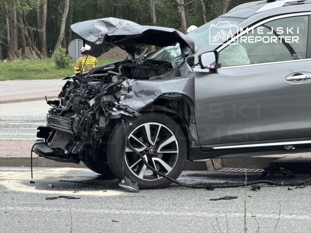 Uszkodzony samochód osobowy z poważnymi zniszczeniami przodu, stojący na drodze w otoczeniu zieleni.