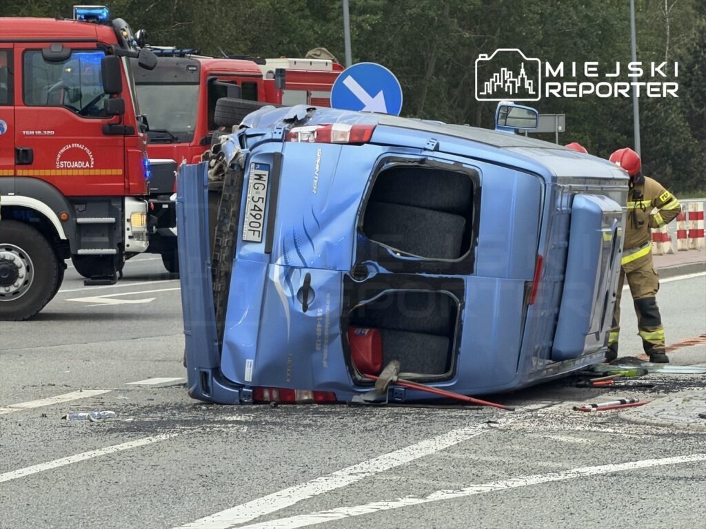 Nieoznakowany niebieski pojazd leży na boku na jezdni, obok pracują strażacy w strojach ochronnych oraz wóz strażacki.