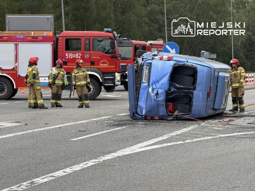Strażacy w mundurach zabezpieczają przewrócony niebieski pojazd na skrzyżowaniu, obok wozu strażackiego.