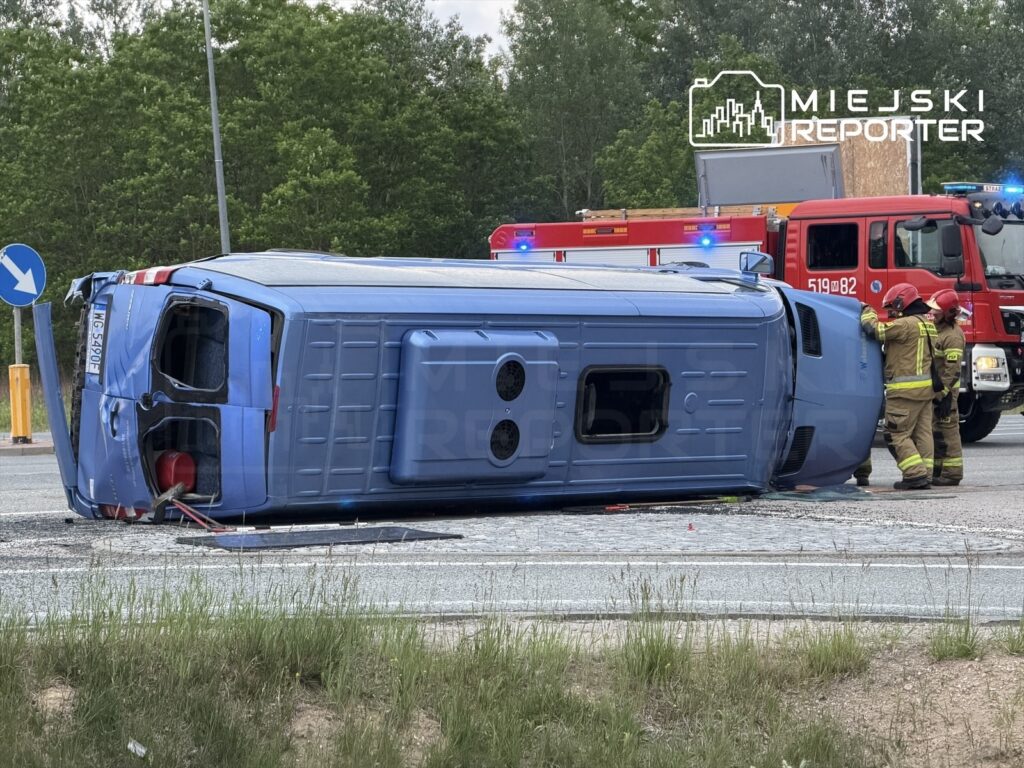 Nieoznakowany niebieski pojazd leży na boku na drodze, obok grupy strażaków w mundurach.