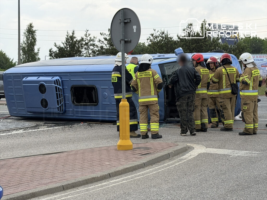 Grupa strażaków w mundurach pomaga osobie przy przewróconym niebieskim pojeździe na skrzyżowaniu.