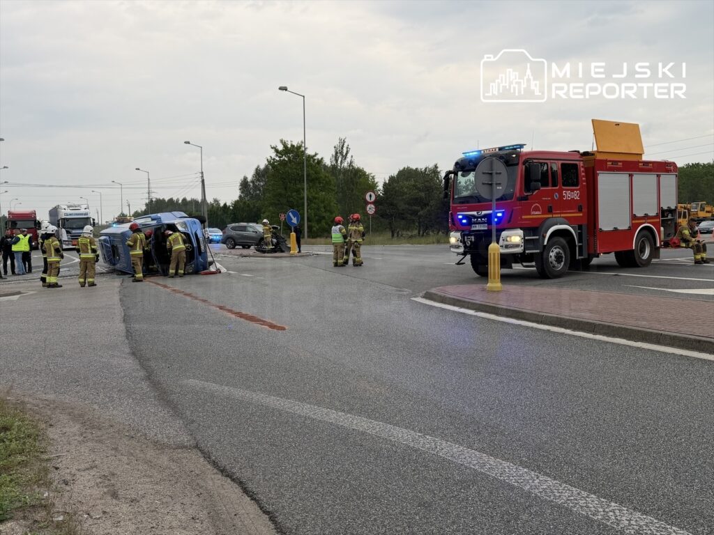 Na drodze leży przewrócony niebieski pojazd, wokół którego pracują strażacy w mundurach. W tle widać woz strażacki z włączonymi sygnałami świetlnymi.