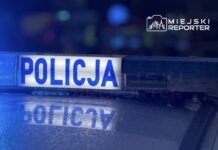 Niebieskie światła policyjne odbijają się na mokrej nawierzchni, z widocznym napisem "POLICJA".