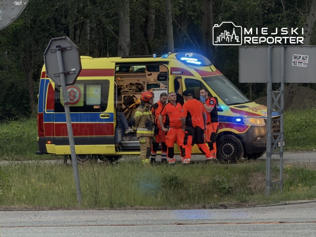 Zespół ratowników w pomarańczowych strojach omawia sytuację obok karetki na drodze w otoczeniu zieleni.