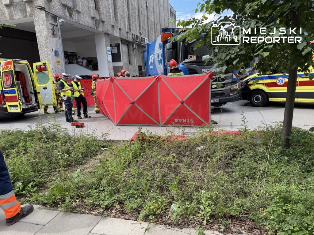 Grupa ratowników w kamizelkach odblaskowych pracuje przy czerwonym namiocie medycznym, obok ambulansów i pojazdów straży pożarnej.