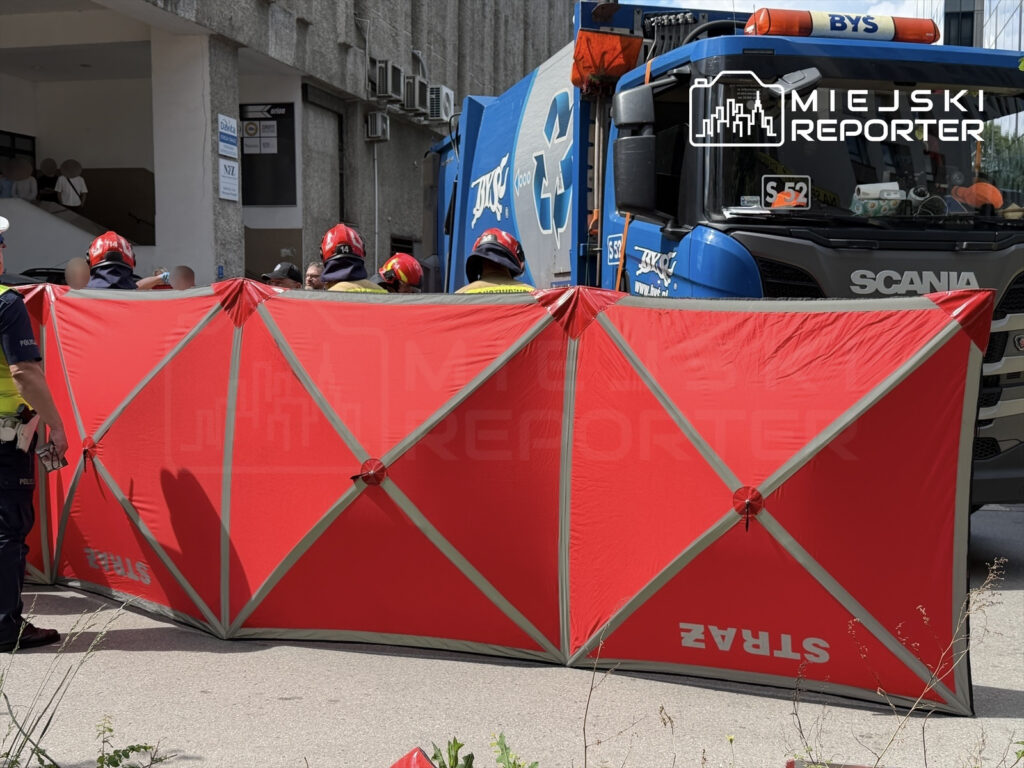 Grupa ratowników w kaskach i odzieży ochronnej ustawia czerwoną osłonę w pobliżu pojazdu służb ratunkowych.