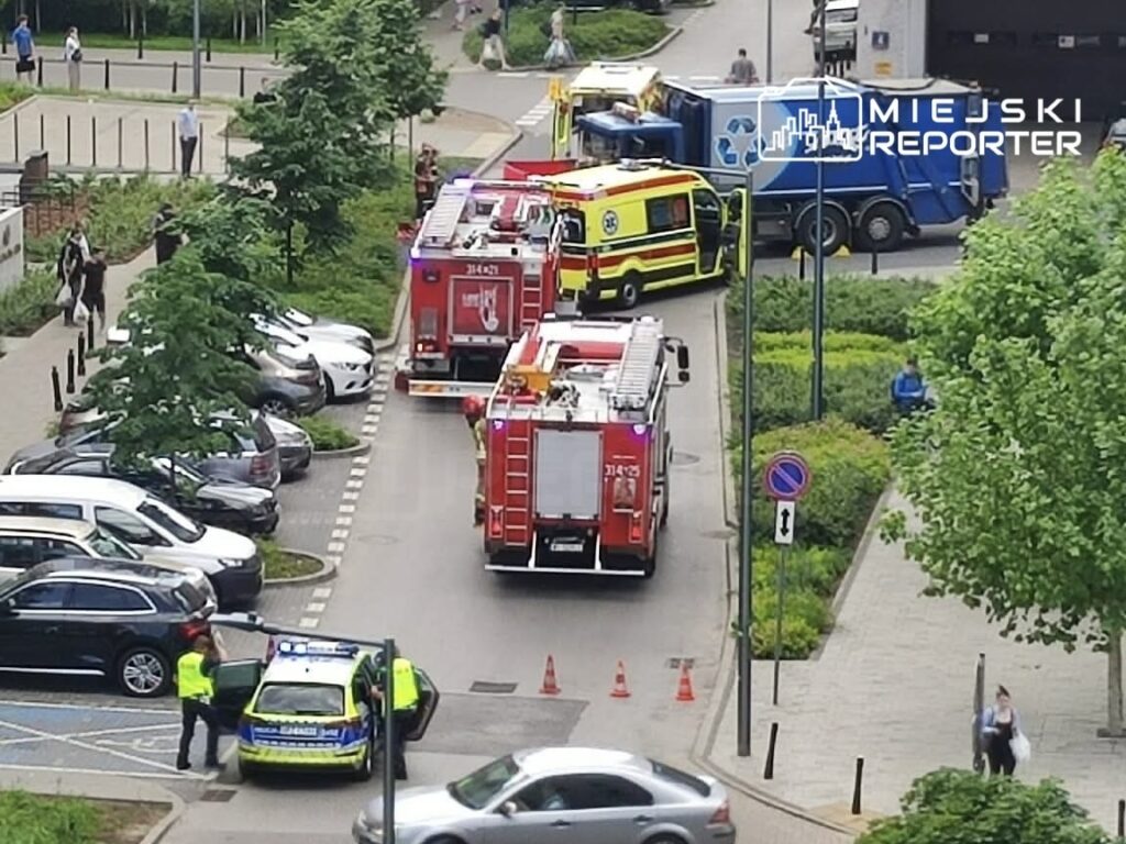 Na parkingu widać pojazdy ratunkowe, w tym straż pożarną i karetkę, oraz policjantów rozmawiających z kierowcami.
