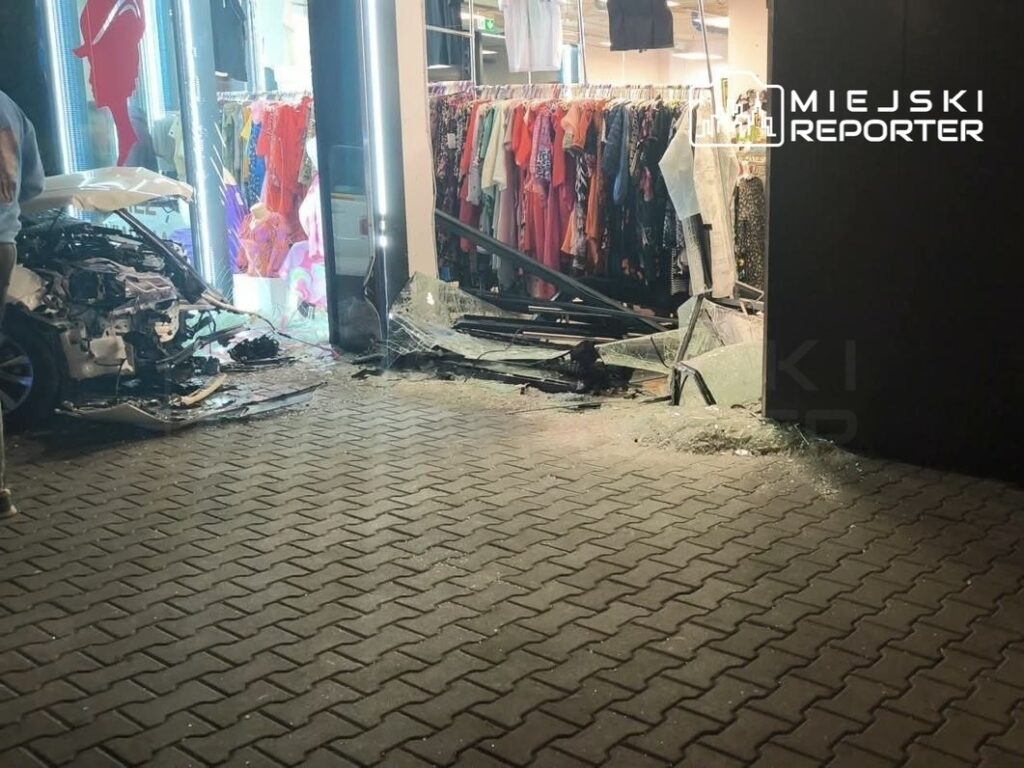 Zniszczony samochód stoi przed witryną sklepu odzieżowego, z rozbitym szkłem i uszkodzeniem ściany.